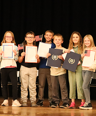  Fourth grade students stand on stage, displaying papers and American flags